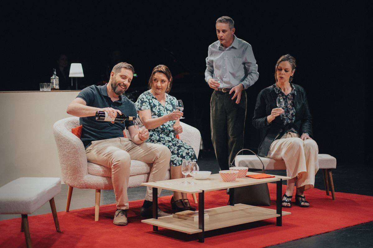 Quatre personnes assises dans un salon moderne, discutant autour d’une table basse, avec un homme servant du vin.
