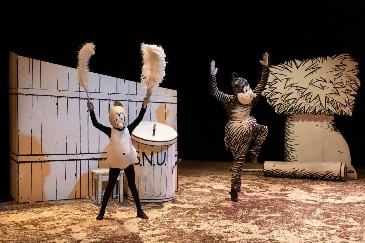Deux personnages costumés sur scène, l’un en zèbre dansant et l’autre en personnage avec des pompons, devant un décor dessiné.