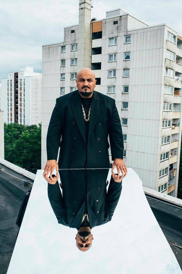 Homme en costume noir debout sur un toit, reflet sur une surface réfléchissante, en arrière-plan des immeubles résidentiels