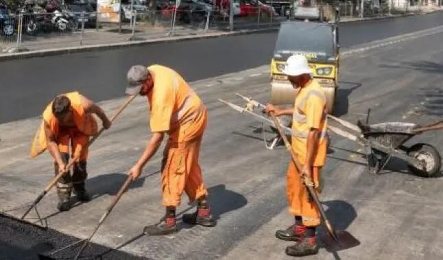 Trois ouvriers en tenue orange travaillent sur la réfection de la chaussée, utilisant des outils pour étaler le goudron.