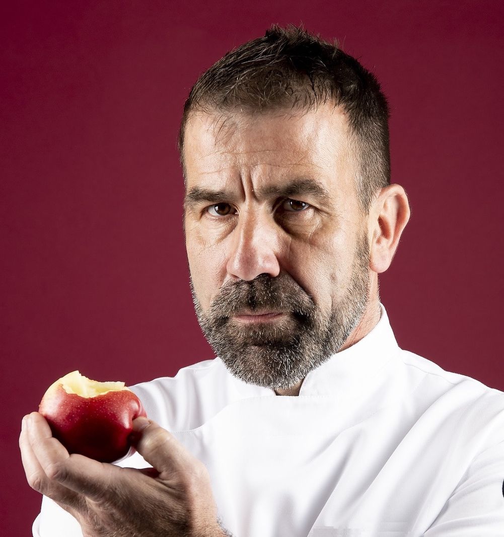 Homme en tenue de chef tenant une pomme croquée devant un fond rouge.