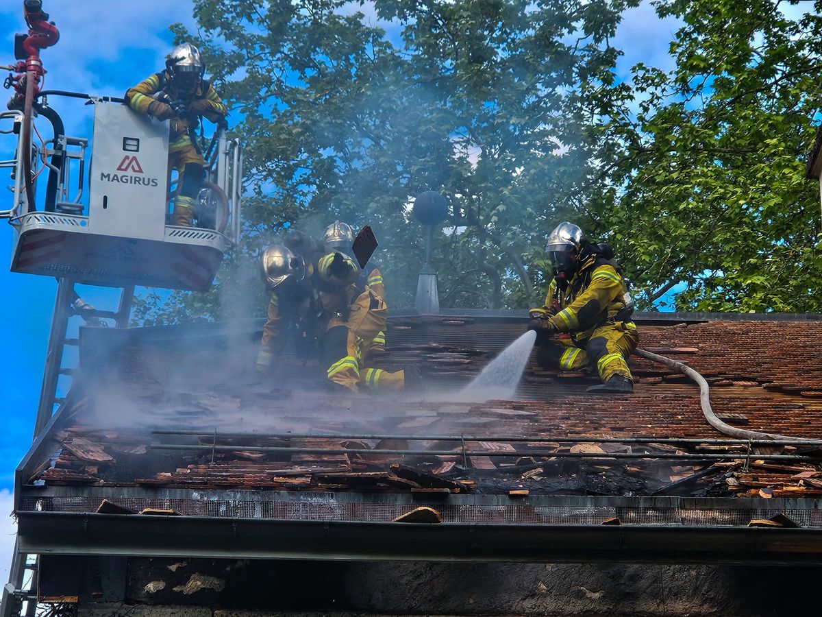 Pompiers en uniforme jaune éteignant un incendie sur un toit avec une lance à incendie, utilisant une plateforme élévatrice, entourés de fumée.