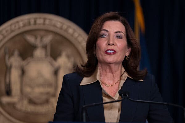 La gouverneure Kathy Hochul, vêtue d’un blazer bleu marine et d’une chemise beige, devant un micro.