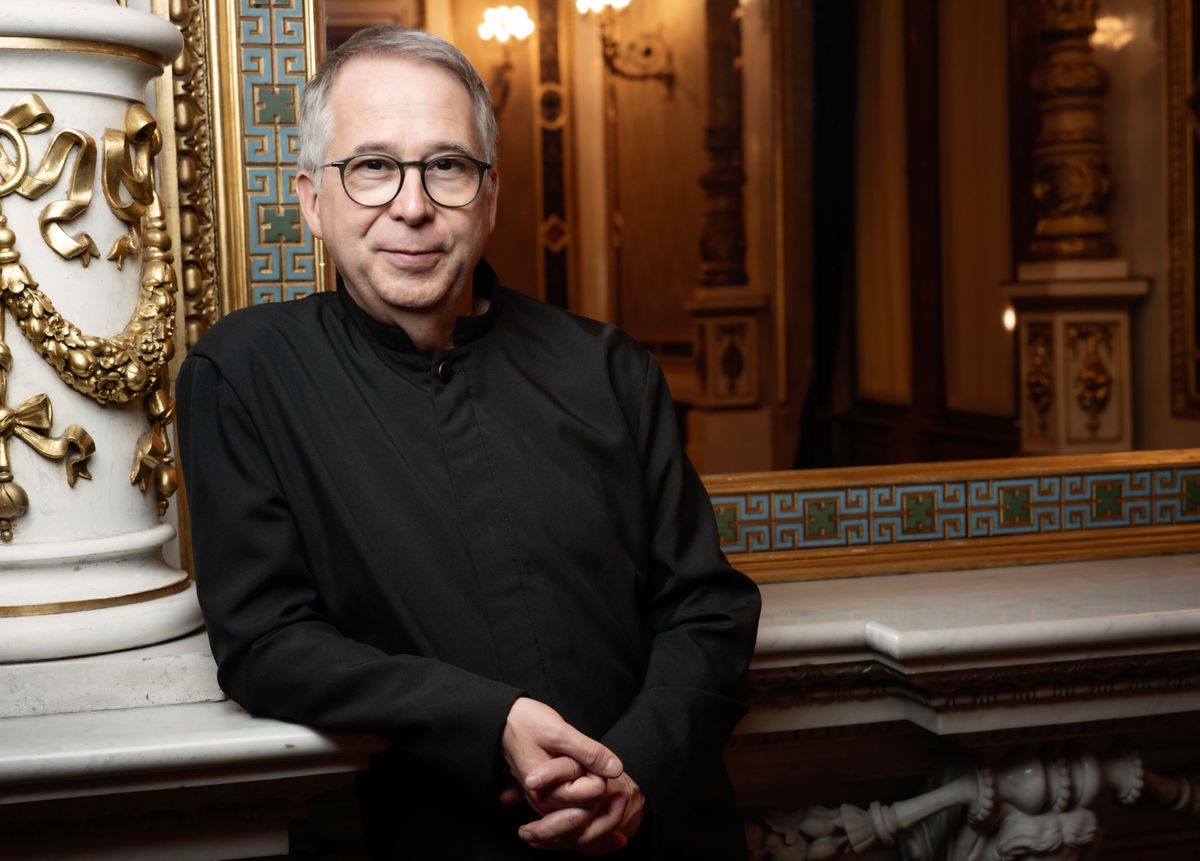 Bertrand de Billy posant dans une salle ornée, portant des lunettes et un costume noir.