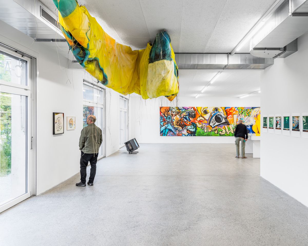 Galerie d’art contemporain avec des œuvres colorées et une installation suspendue. Deux personnes observent les œuvres exposées.