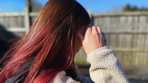 Profil de Corinna avec les cheveux rouges, regardant le sol et se protégeant le visage avec la main droite