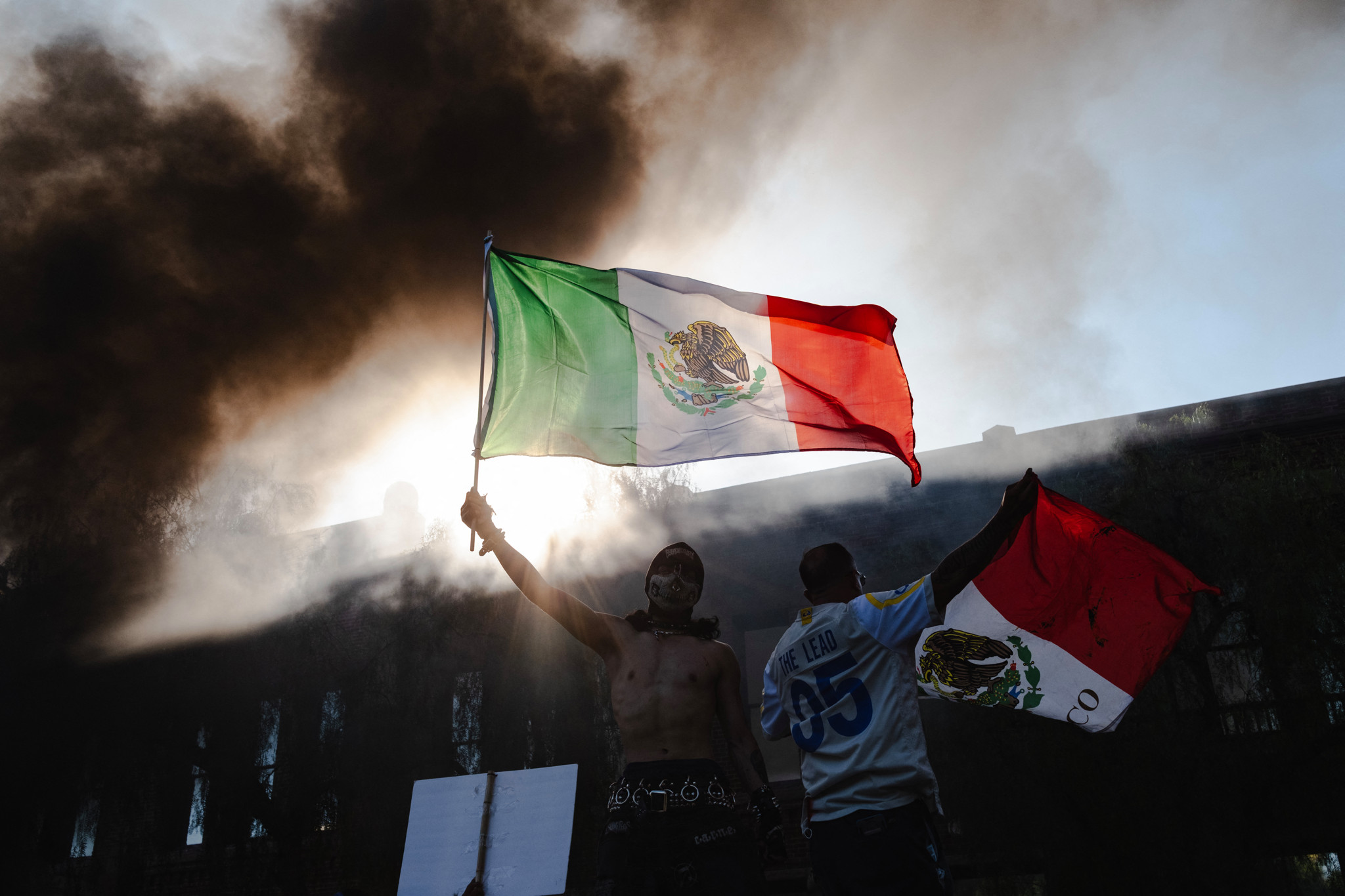 Un manifestant à Los Angeles tenant un drapeau mexicain devant des voitures en flammes après des opérations d'ICE.