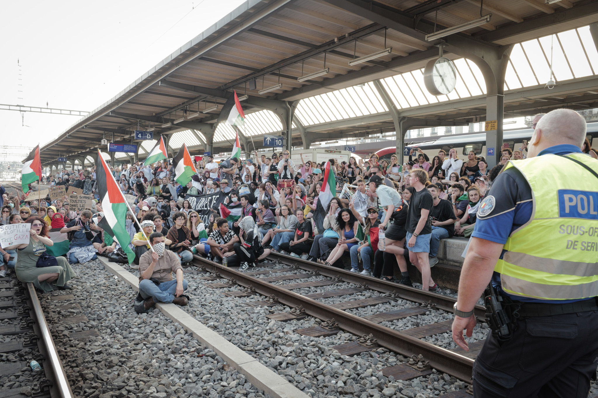 Manifestants assis sur les voies de la gare Cornavin, à Genève, brandissant des drapeaux palestiniens lors d’une protestation contre la guerre à Gaza, confrontés à un policier.