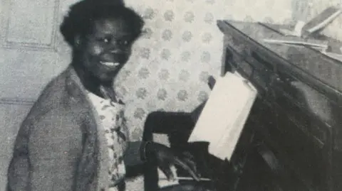 Grace Adebowale dans les années 40, souriant à la caméra tout en jouant du piano.