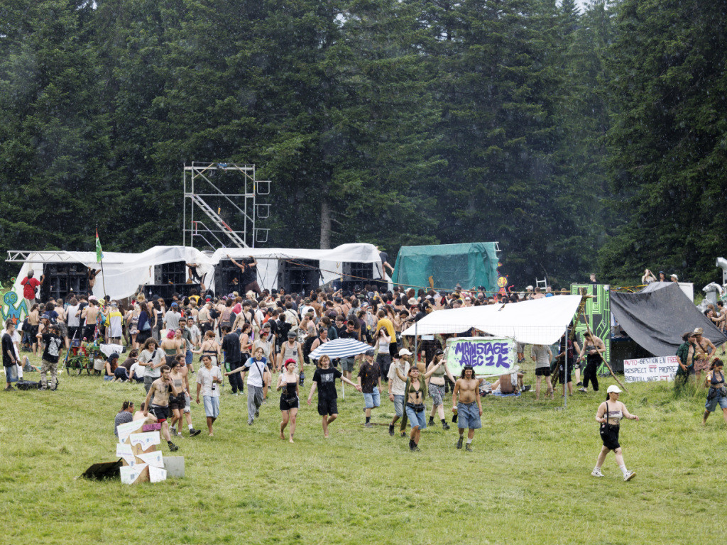 Participants quittant une rave party illégale en plein air à Malleray, entourés de tentes et d’équipements de sonorisation, avec une forêt en arrière-plan.