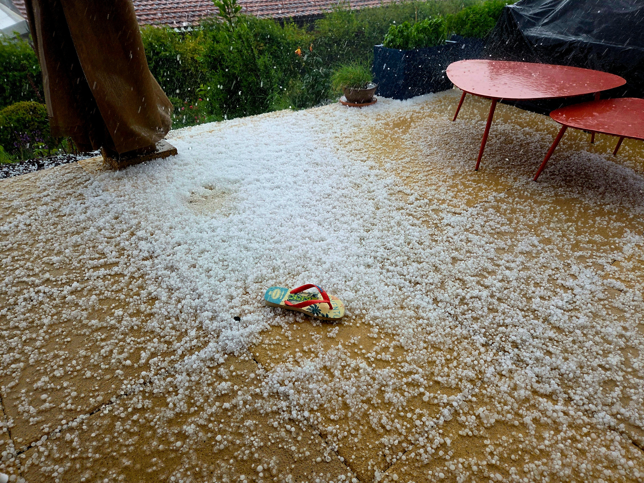 Une violente tempête de grêle couvre une terrasse extérieure avec un mélange de grêle et de neige. Une unique sandale est laissée par terre, entourée par de la grêle, tandis que deux tables rouges sont visibles sur le côté droit.