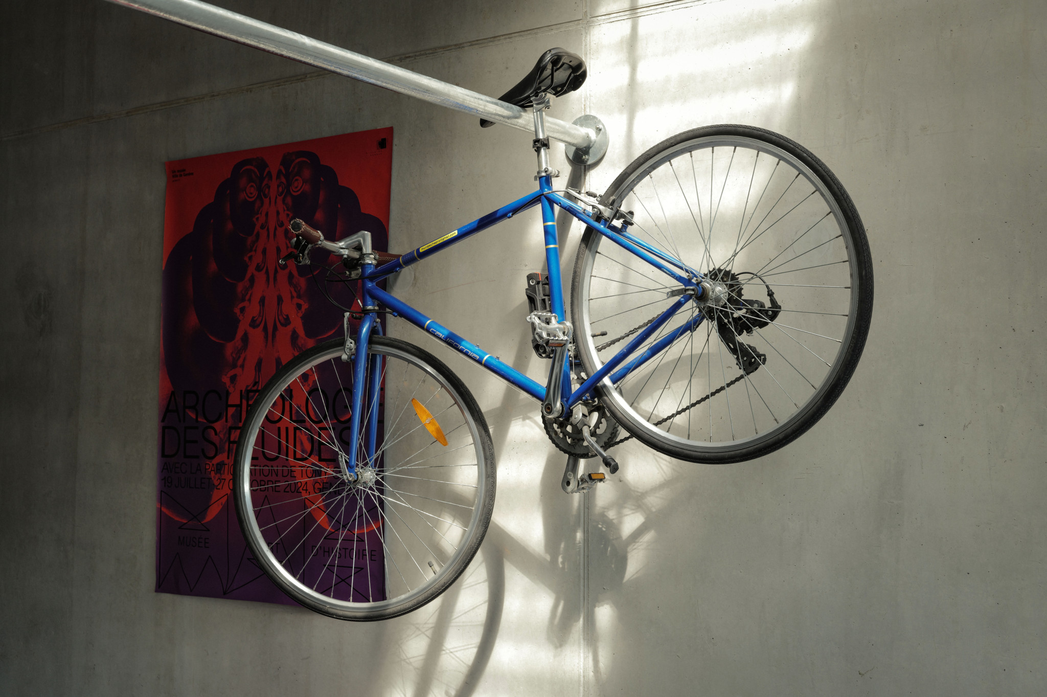 Un vélo bleu suspendu verticalement à un mur en béton, à côté d’une affiche colorée de l’exposition ’Architecture des Guides’ à Genève.