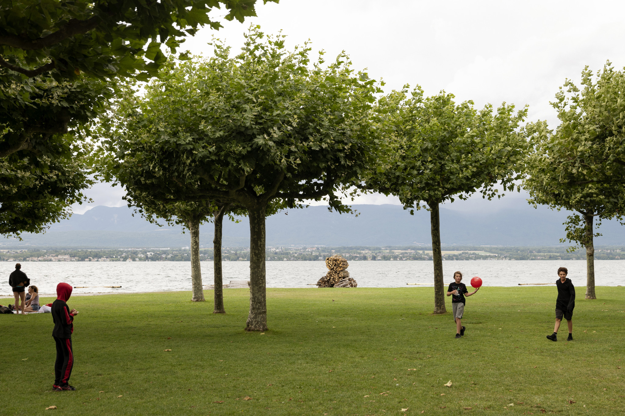 Des enfants jouent avec un ballon rouge sur une pelouse près du lac, entourés d’arbres, à Hermance, un village à la frontière franco-suisse, un jour de 1ᵉʳ août.