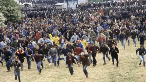 Des hommes courant sur un grand terrain, avec des policiers en arrière-plan.