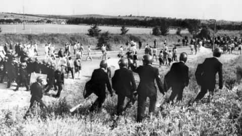 Image en noir et blanc de mineurs et de policiers en tenue anti-émeute.