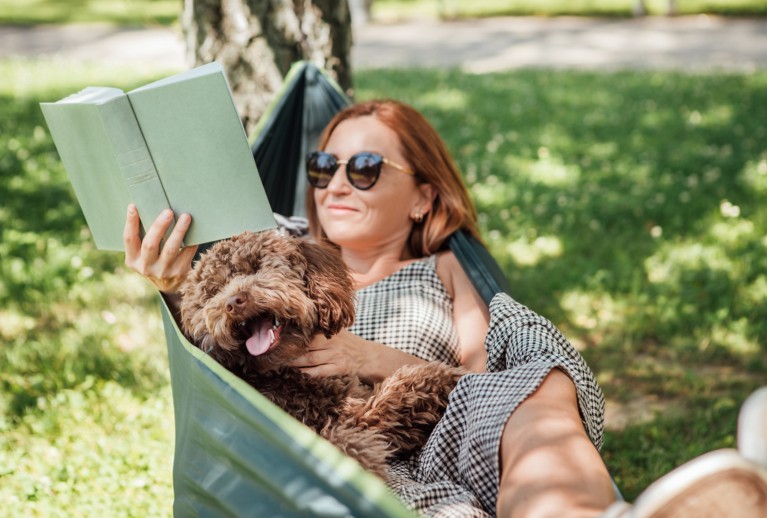 Une femme aux cheveux roux lisant un livre dans un hamac avec son chien.