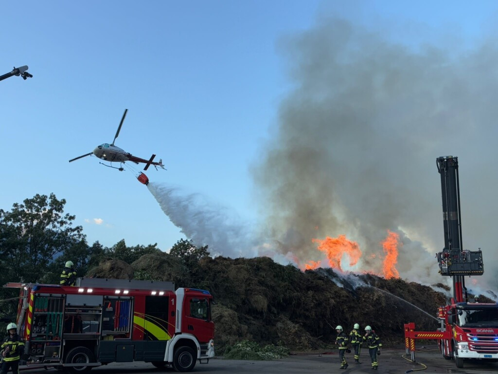 Un hélicoptère d’Air Glaciers largue de l’eau sur un incendie, tandis que des pompiers luttent contre les flammes au sol.