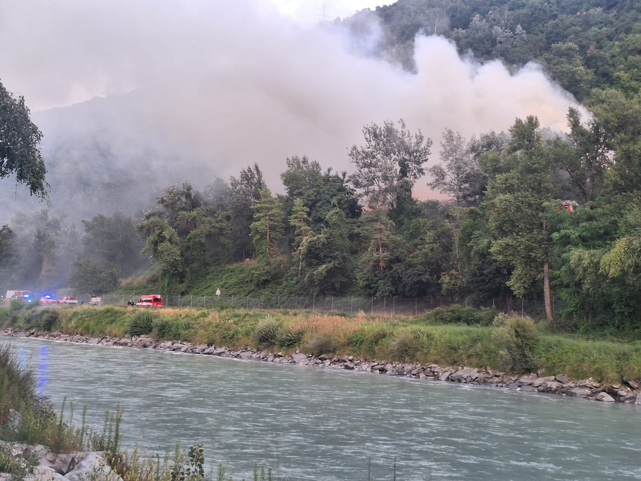 Feu de forêt sur une colline avec fumée dense, voitures de pompiers visibles près d’une rivière, végétation luxuriante.